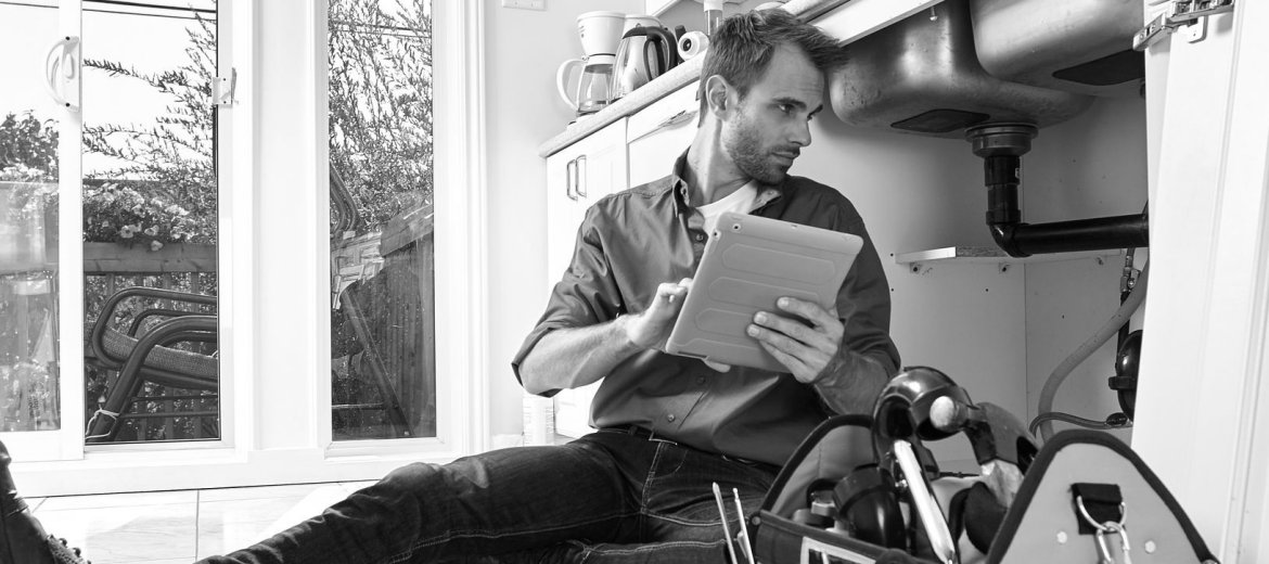 Man inspecting plumbing under a sink, with sash windows in the background.